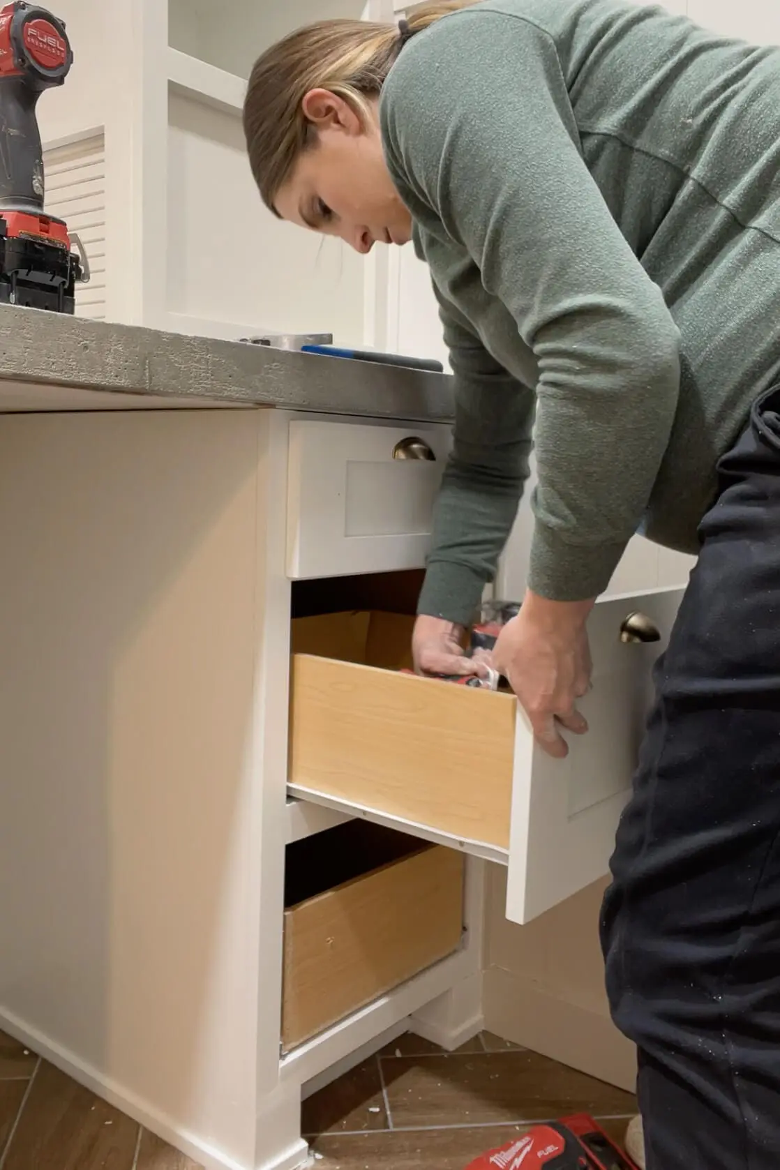 Person adjusting and pulling open a white bathroom cabinet drawer to test alignment and fit during a renovation. The concrete countertop and installed shelving unit are visible above.